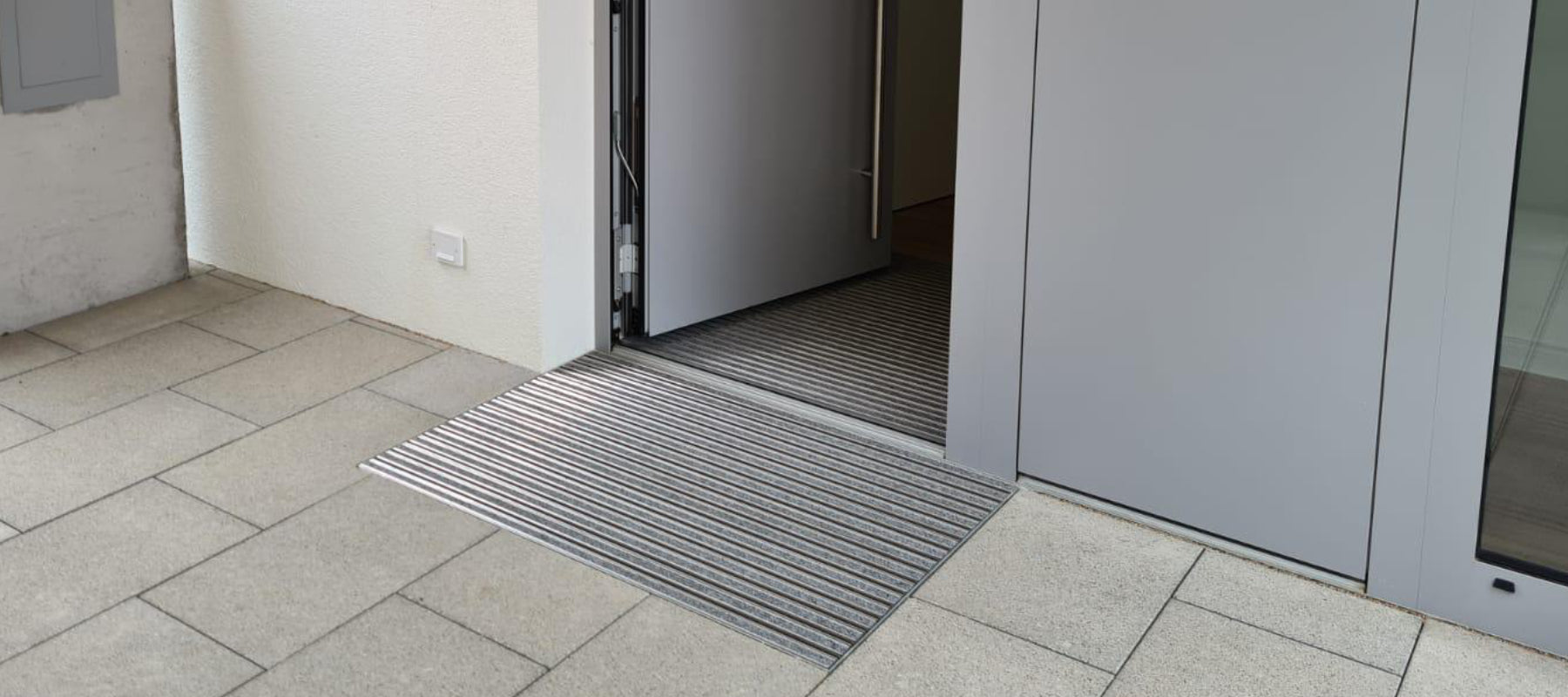 Vor einer grauen Haustür liegt eine Fußmatte eingelassen in Bodenfliesen