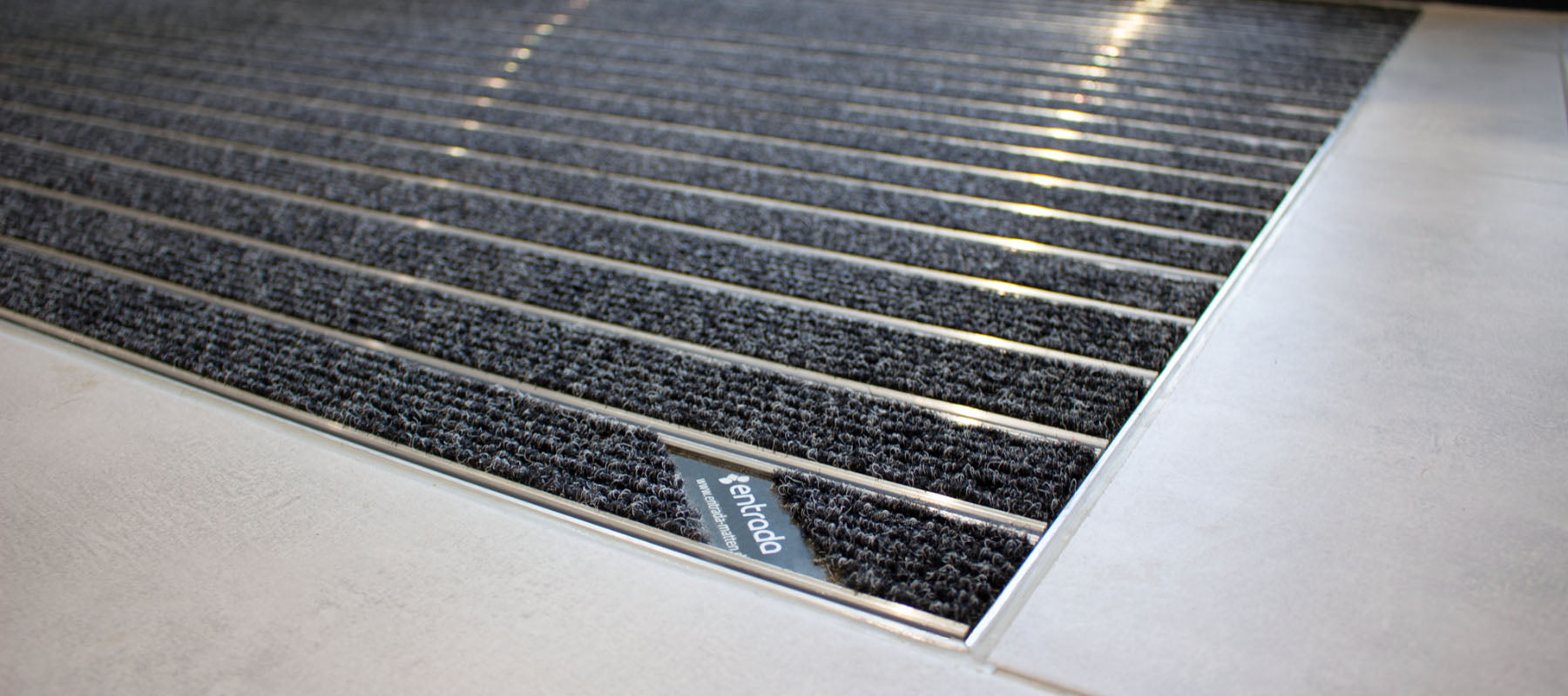 Aluprofilmatte mit entrada-Logo liegt in Bodenvertiefung eines grauen Fliesenbodens