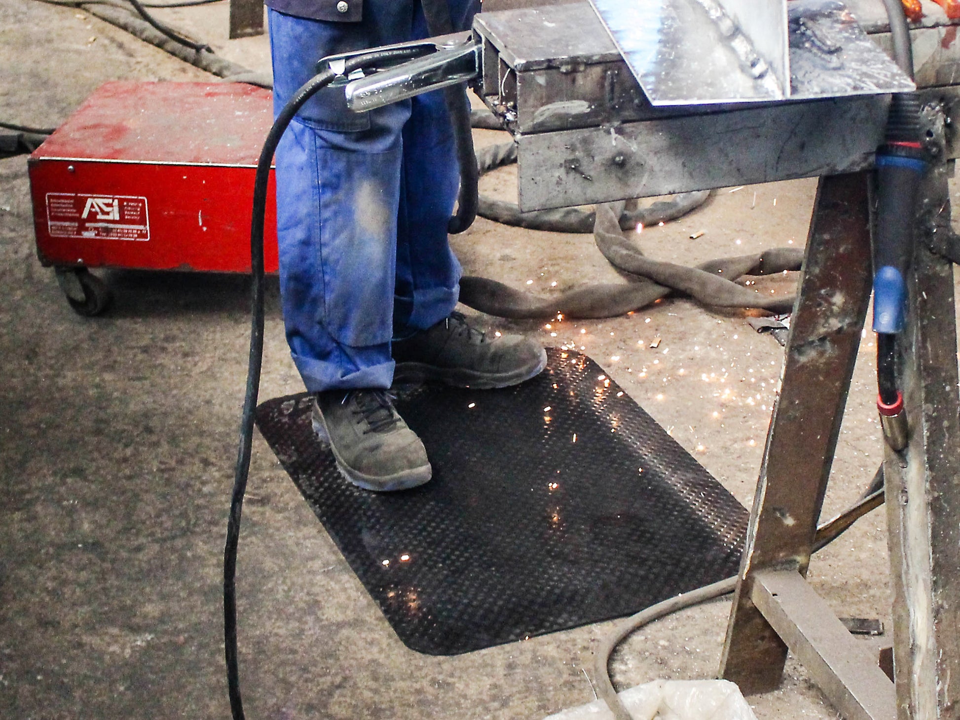Arbeiter steht auf ergonomischer Steharbeitsplatzmatte in Industriehalle, Funken fliegen durch die Luft