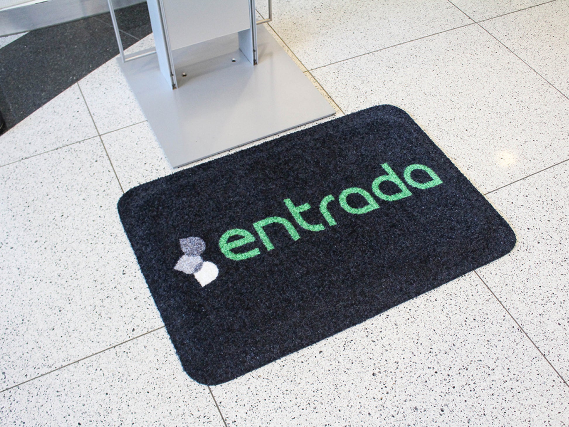 Arbeitsplatzmatte mit Teppichoberfläche mit Aufdruck entrada liegt auf einem weißen Fliesenboden