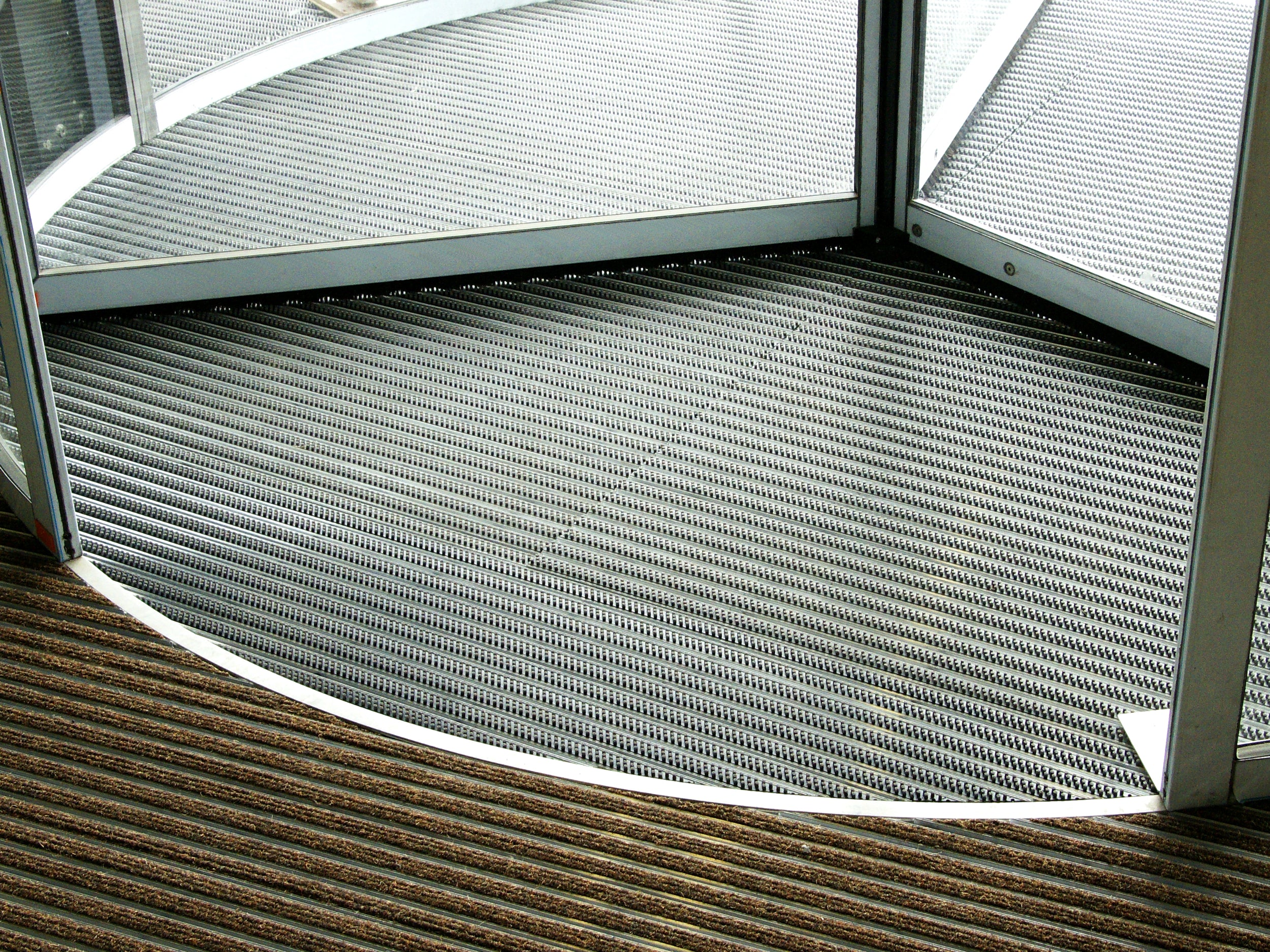 Fußabtreter aus Aluminium mit Rips- und Bürsteneinsatz in rund liegt in Drehtür mit Abdeckschiene aus Edelstahl