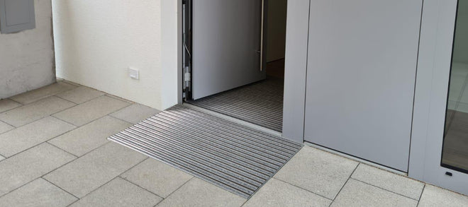 Vor einer grauen Haustür liegt eine Fußmatte eingelassen in Bodenfliesen