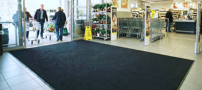 In einem Supermarkt Eingangsbereich laufen Personen mit Einkaufswagen- und taschen durch die Eingangstür. Im Eingangsbereich liegt eine große Schmutzfangmatte und es stehen Pflanzen und Einkaufswagen.