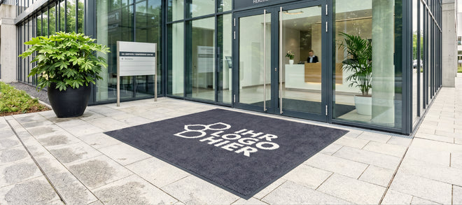 Große schwarze Logomatte mit der Aufschrift "Ihr Logo hier" liegt außen vor einer Glastür
