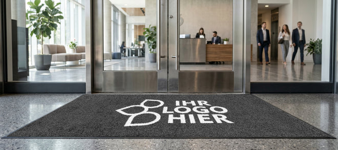 Eine graumelierte Fußmatte mit dem Aufdruck "Ihr Logo hier" liegt vor der Glastür eines Bürogebäudes.