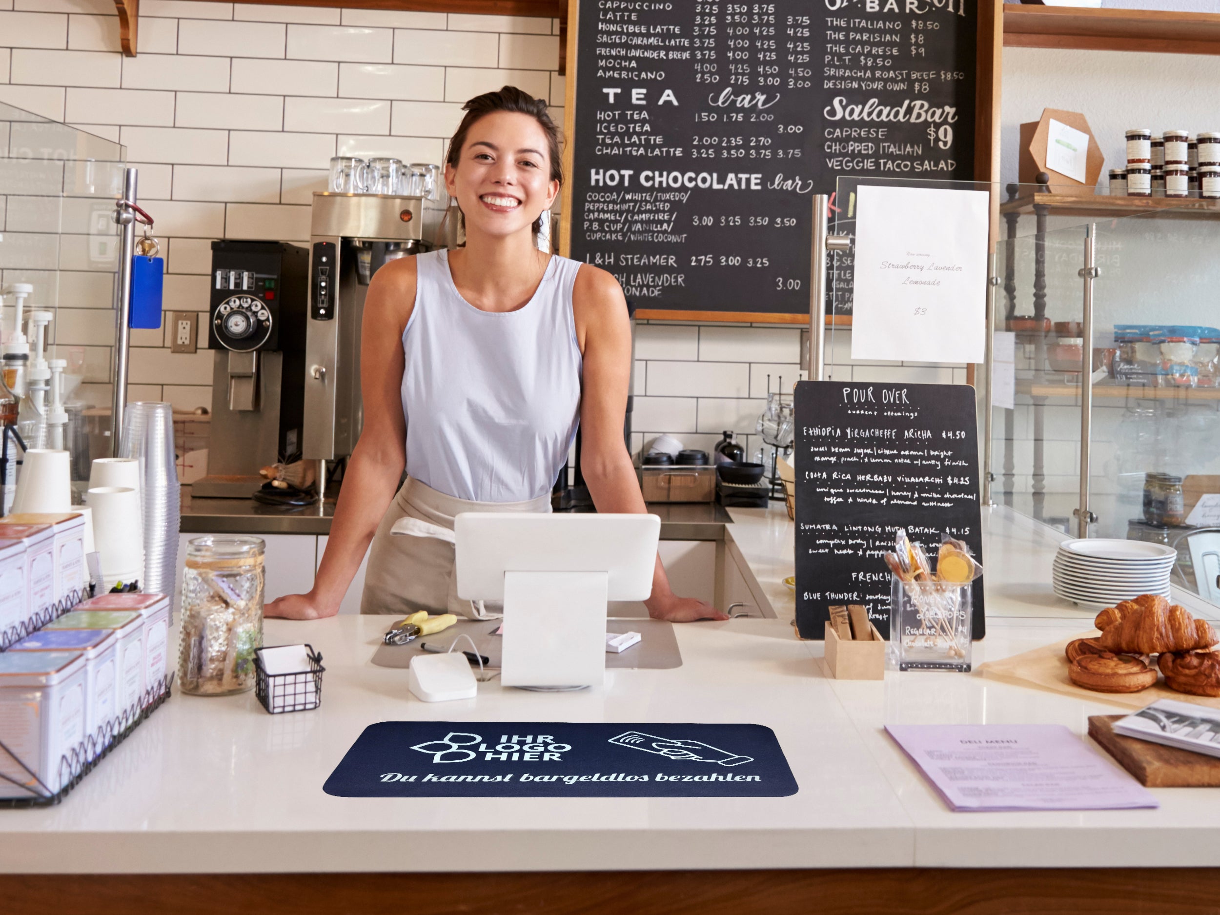 In einem Café liegt eine Thekenmatte auf dem Tresen mit Schriftzug: Du kannst bargeldlos bezahlen. Dahinter steht eine junge lachende Frau.